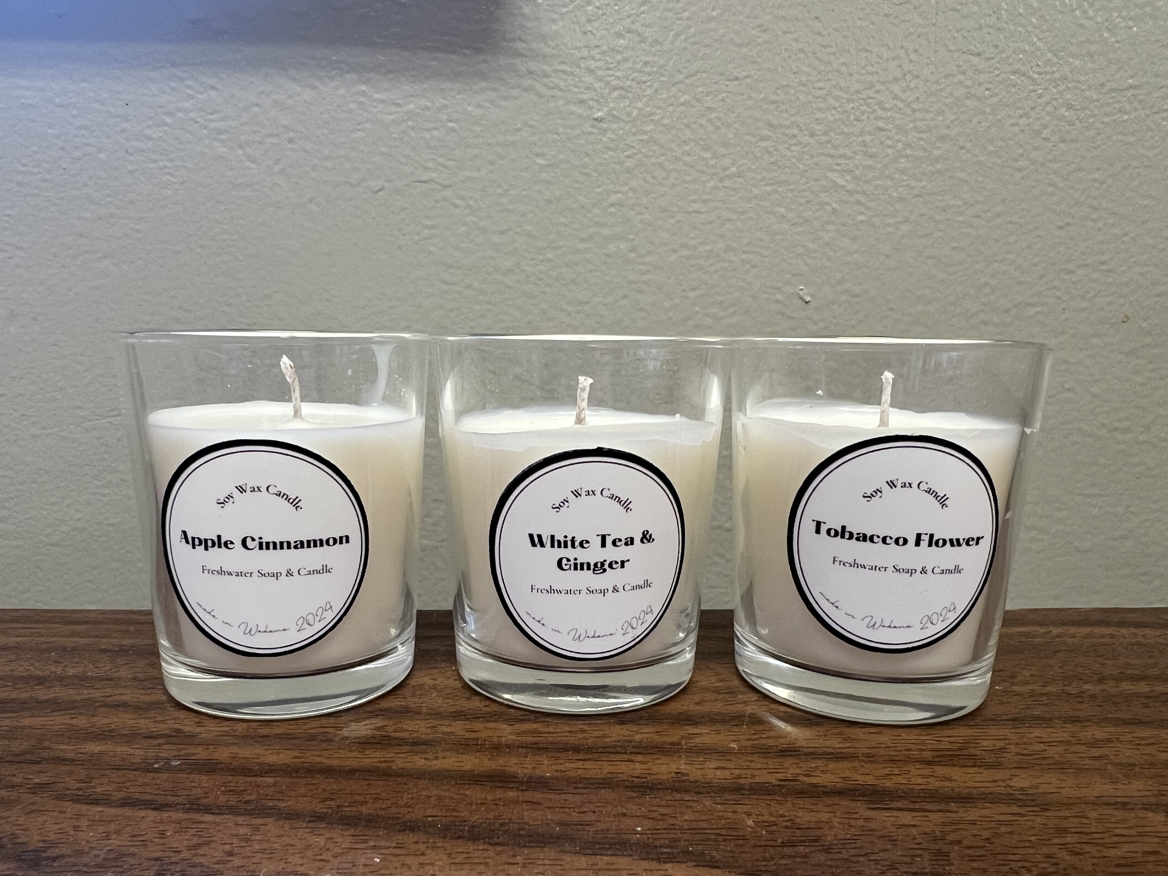 Three glass candles with labels displaying "Apple Cinnamon", "White Tea &amp; Ginger", and "Tobacco Flower" are placed on a wooden surface against a plain white background.