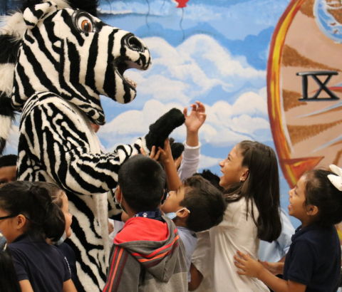 A group of children enthusiastically interacting with a large zebra-patterned figure against a backdrop of a painted sky and clock.