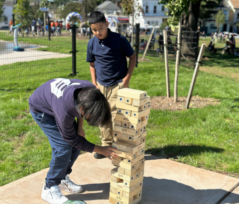 Two people, a man and a woman, are playing a game of Jenga on a grassy area in an outdoor setting with trees and buildings visible in the background.