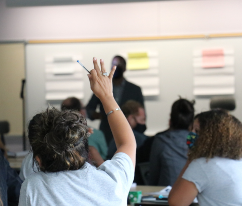 A classroom setting with students raising their hands, while a person stands in the front of the room, likely a teacher or instructor.