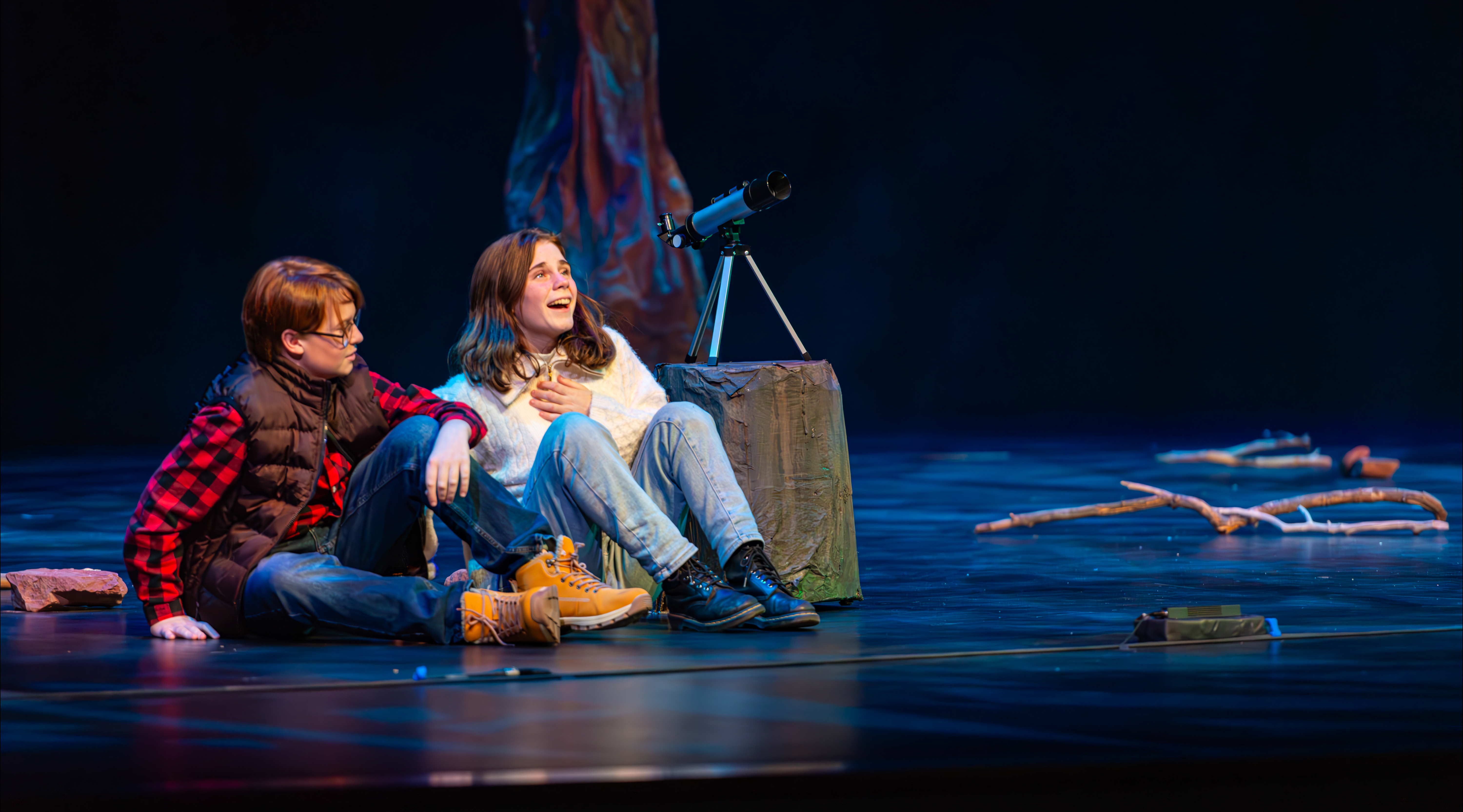 two middle school students acting out a scene on a theater with a telescope behind them
