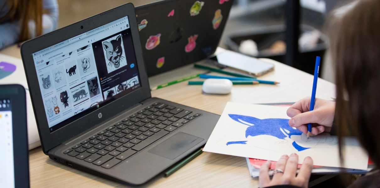 A laptop displaying an online interface, with a person's hands drawing a blue butterfly on a piece of paper in the foreground.