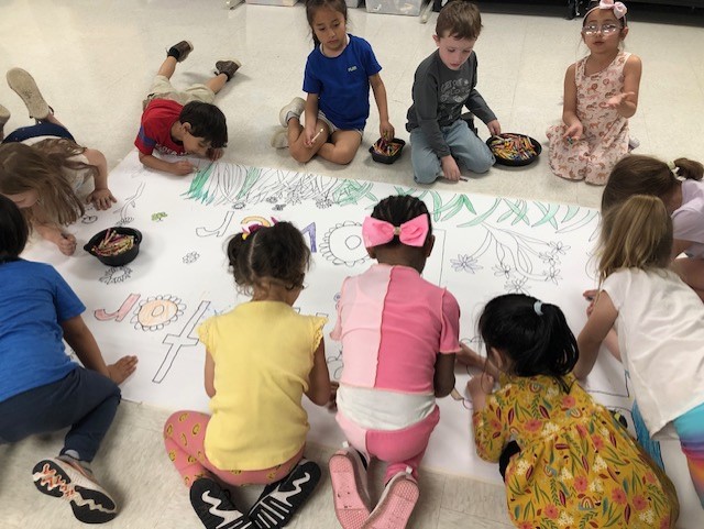 A group of children sitting on the floor, engaged in a collaborative drawing activity, with various colorful drawings visible on the large sheet of paper in front of them.