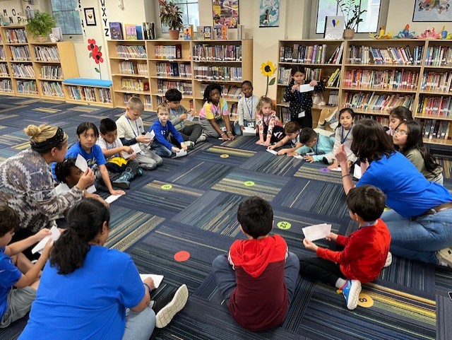 A group of children sitting on the floor in a library, surrounded by bookshelves and various decorations on the walls.