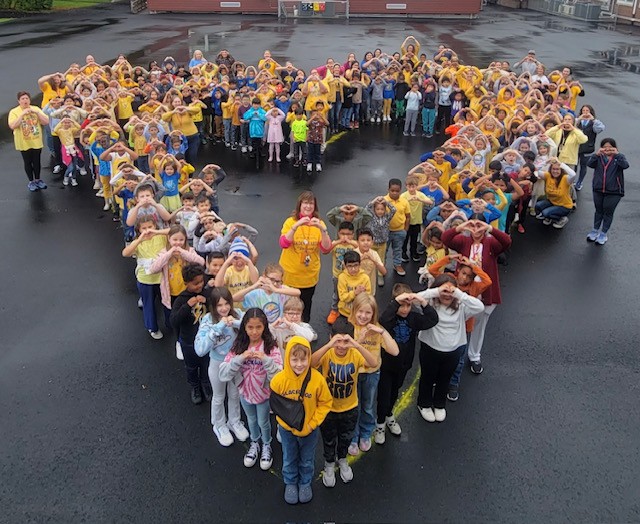 A large group of people, mostly wearing yellow shirts, are gathered in the shape of a heart on a paved outdoor area, with buildings visible in the background.