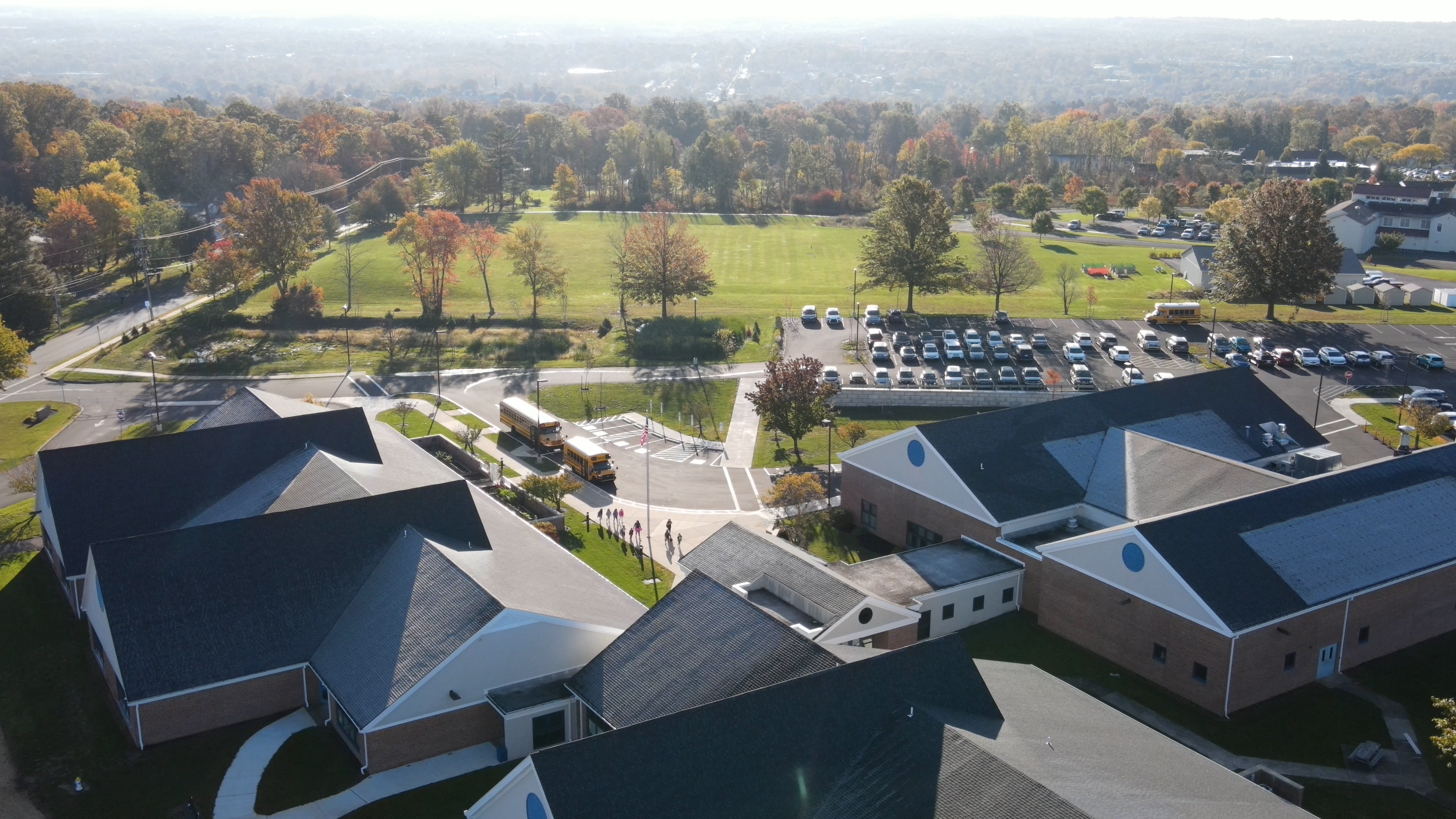 A scenic aerial view of a quaint village nestled among lush, autumnal forests, with a grassy field in the foreground and a parking lot visible in the distance.