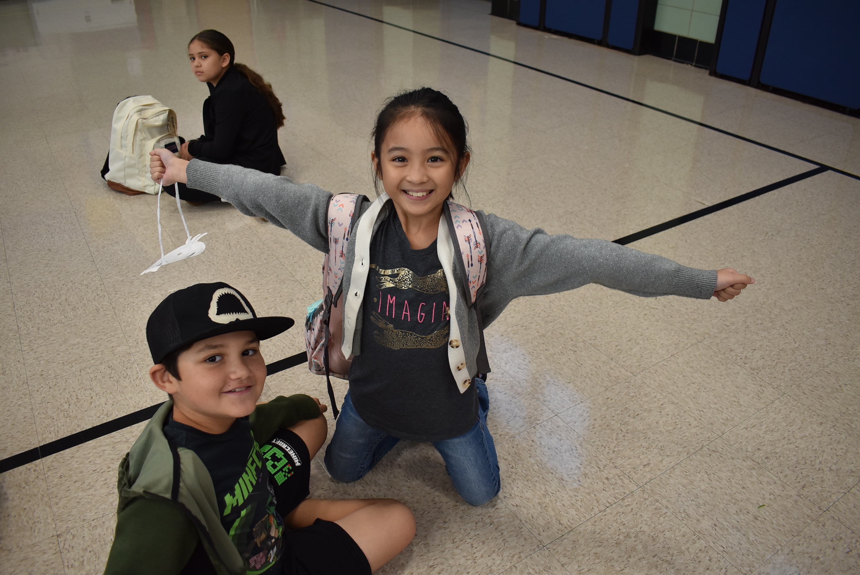 Two young people, a boy and a girl, are standing in a hallway. The girl is smiling and appears to be in a playful mood, while the boy is sitting on the floor.