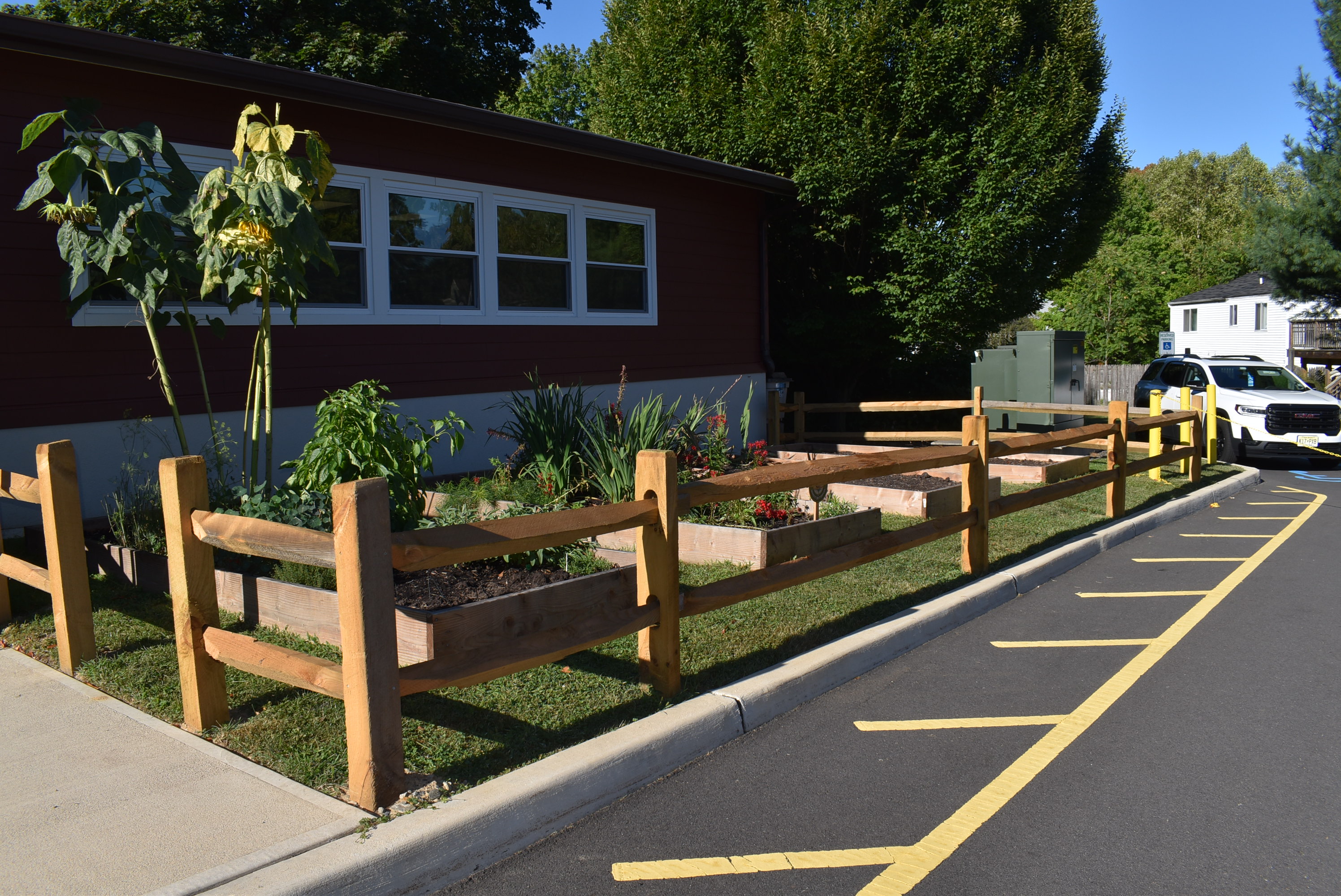 A wooden fence surrounds raised garden beds in the foreground, with a red building and lush greenery in the background.