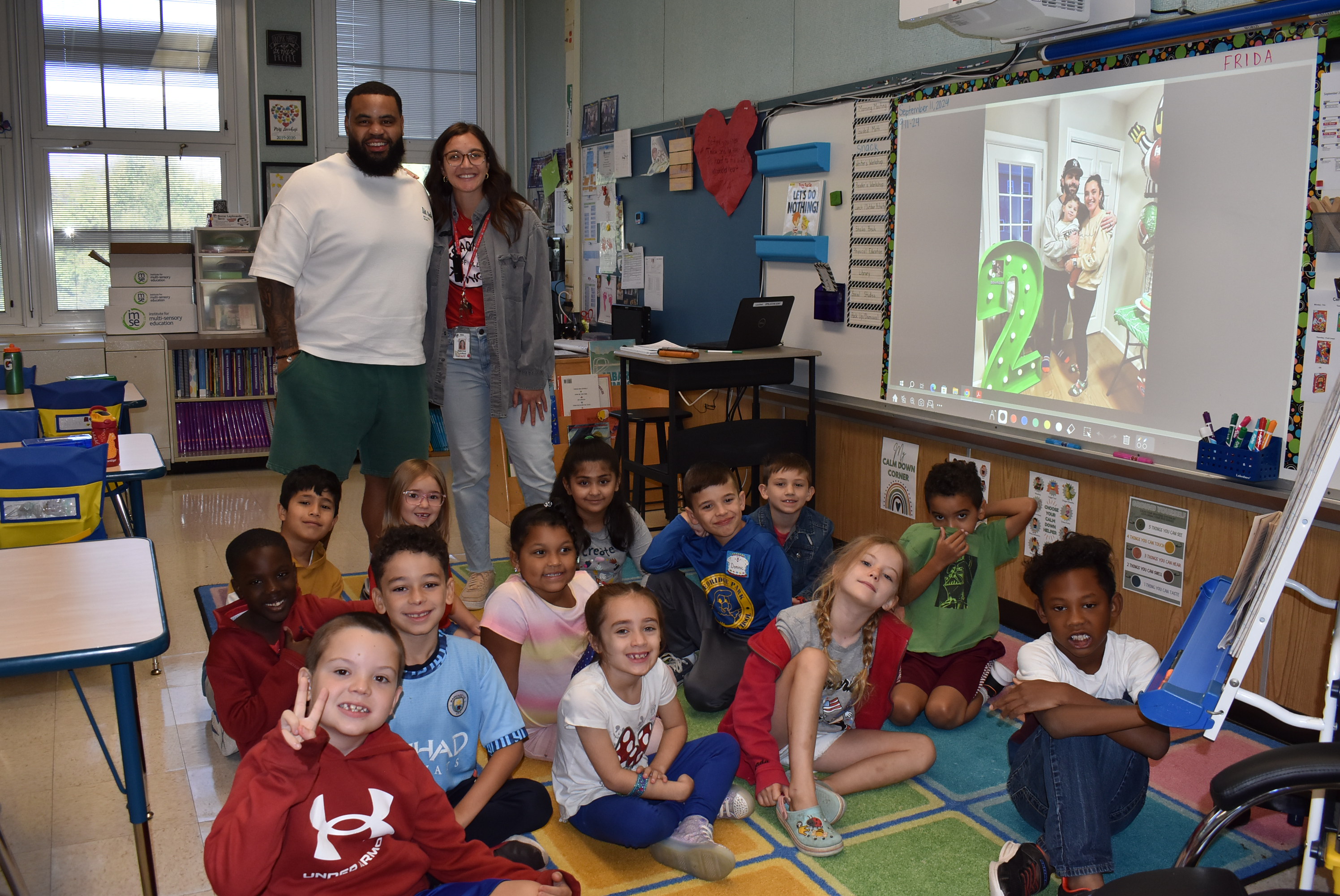 The image shows a group of young students sitting on the floor of a classroom, with a teacher and another adult standing behind them. The classroom appears to be well-equipped with various educational materials and displays on the walls.