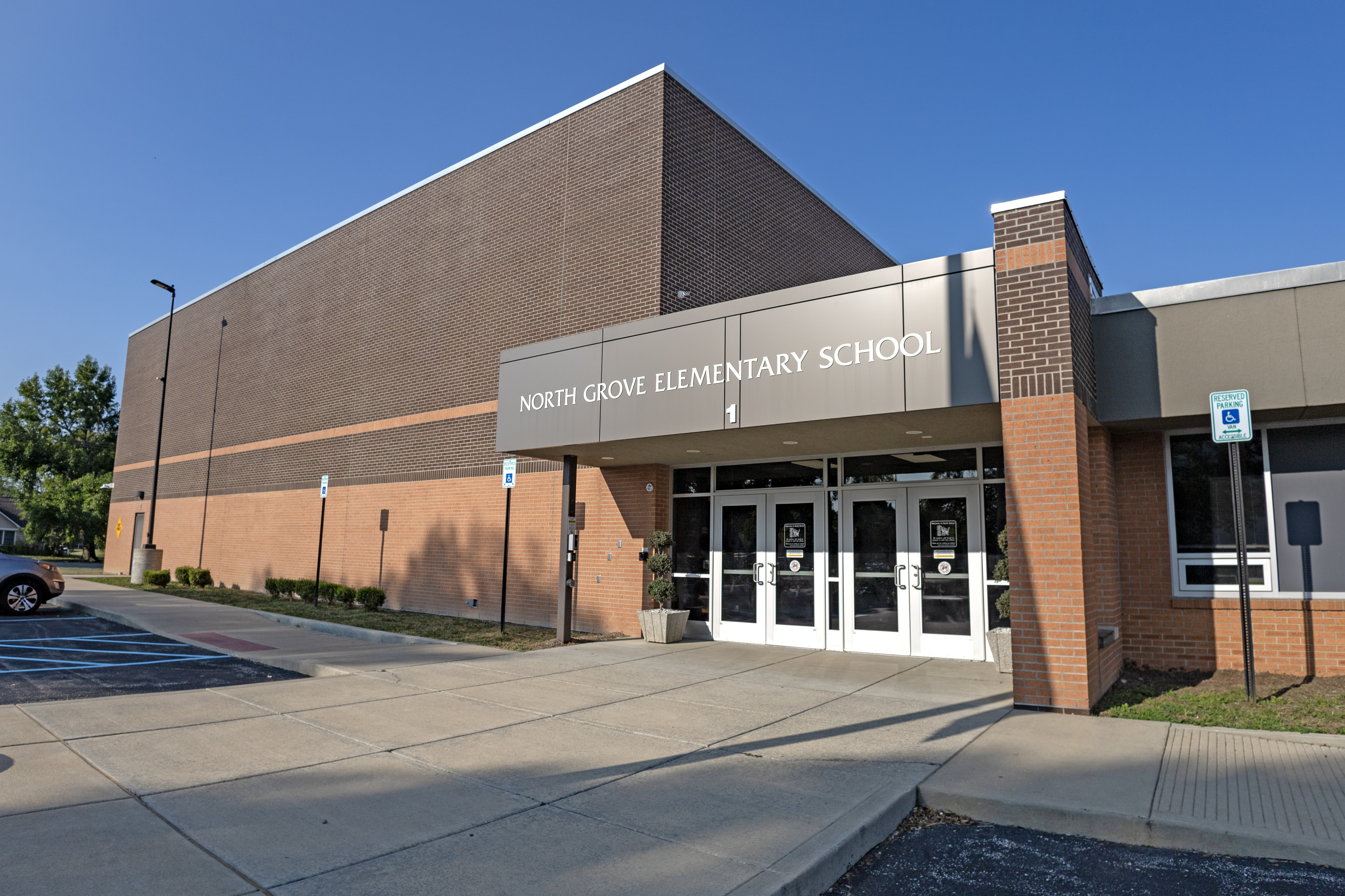 A modern brick building labeled "North Grove Elementary School" stands under a clear blue sky, with a paved walkway and parking lot in the foreground.