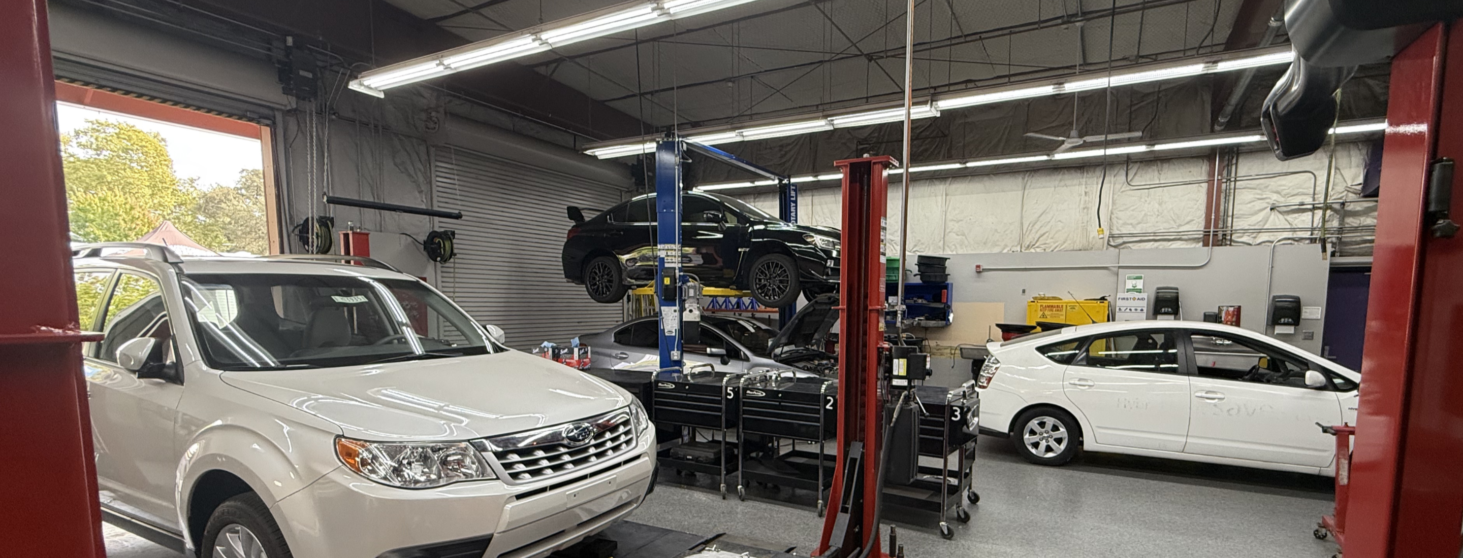The image shows a garage or auto repair shop with several vehicles, including a white SUV in the foreground and a black car on a lift in the background.