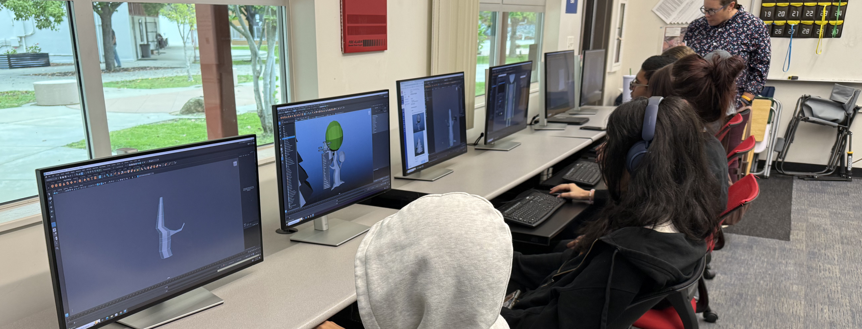 The image shows a computer lab with several people seated at desks, working on computers that display various graphical interfaces.