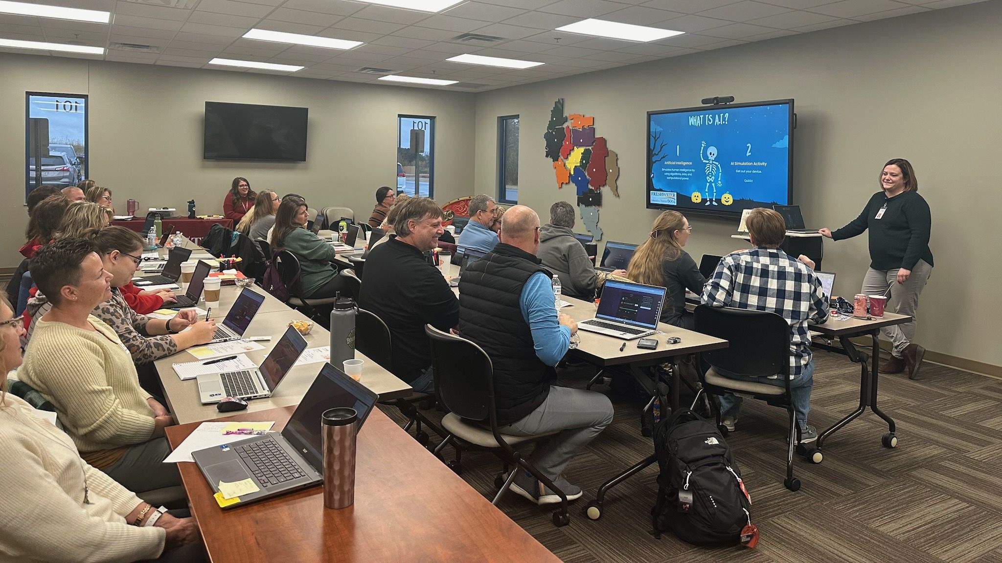A classroom-like setting with several people seated at desks, using laptops and computers, while a presenter stands in front of a large display screen.