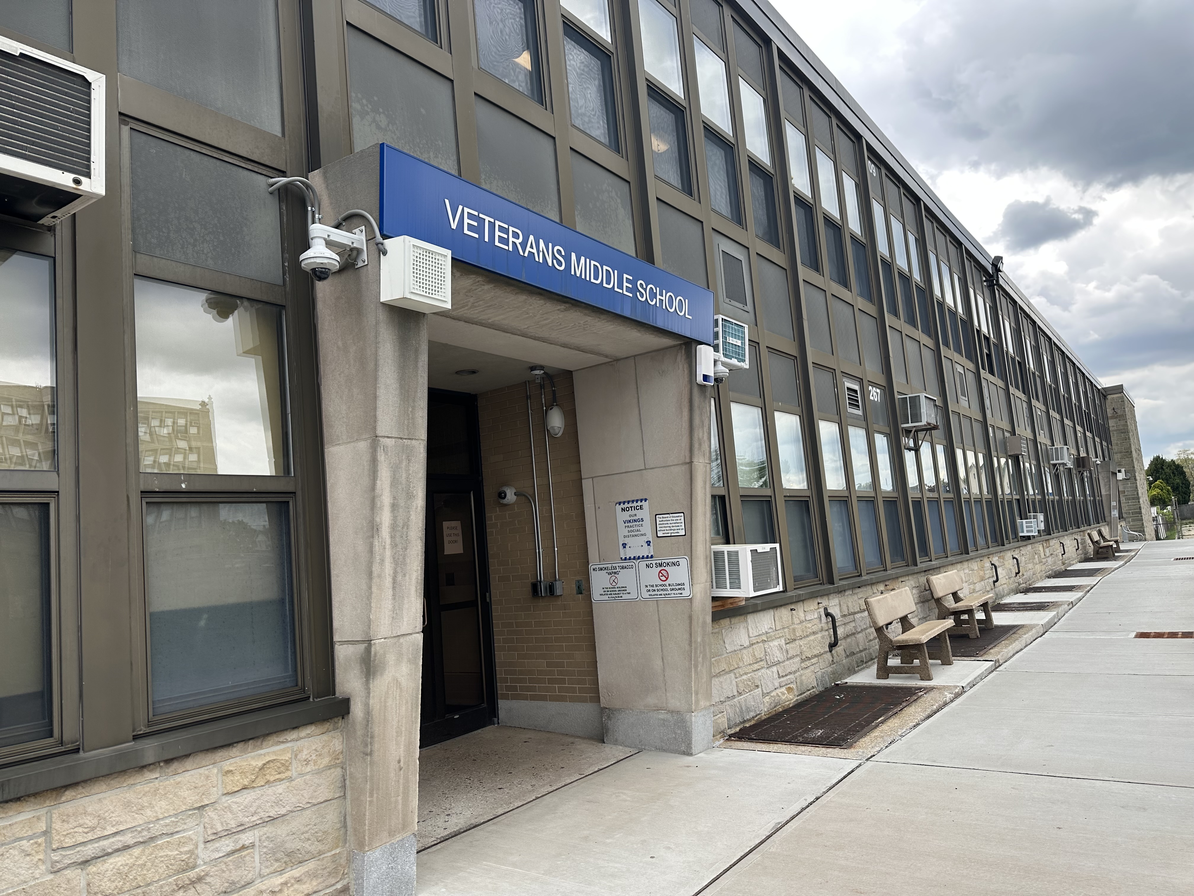 A concrete walkway leads to the entrance of Veterans Middle School, with a long, multi-story brick and stone building stretching into the background under a cloudy sky.