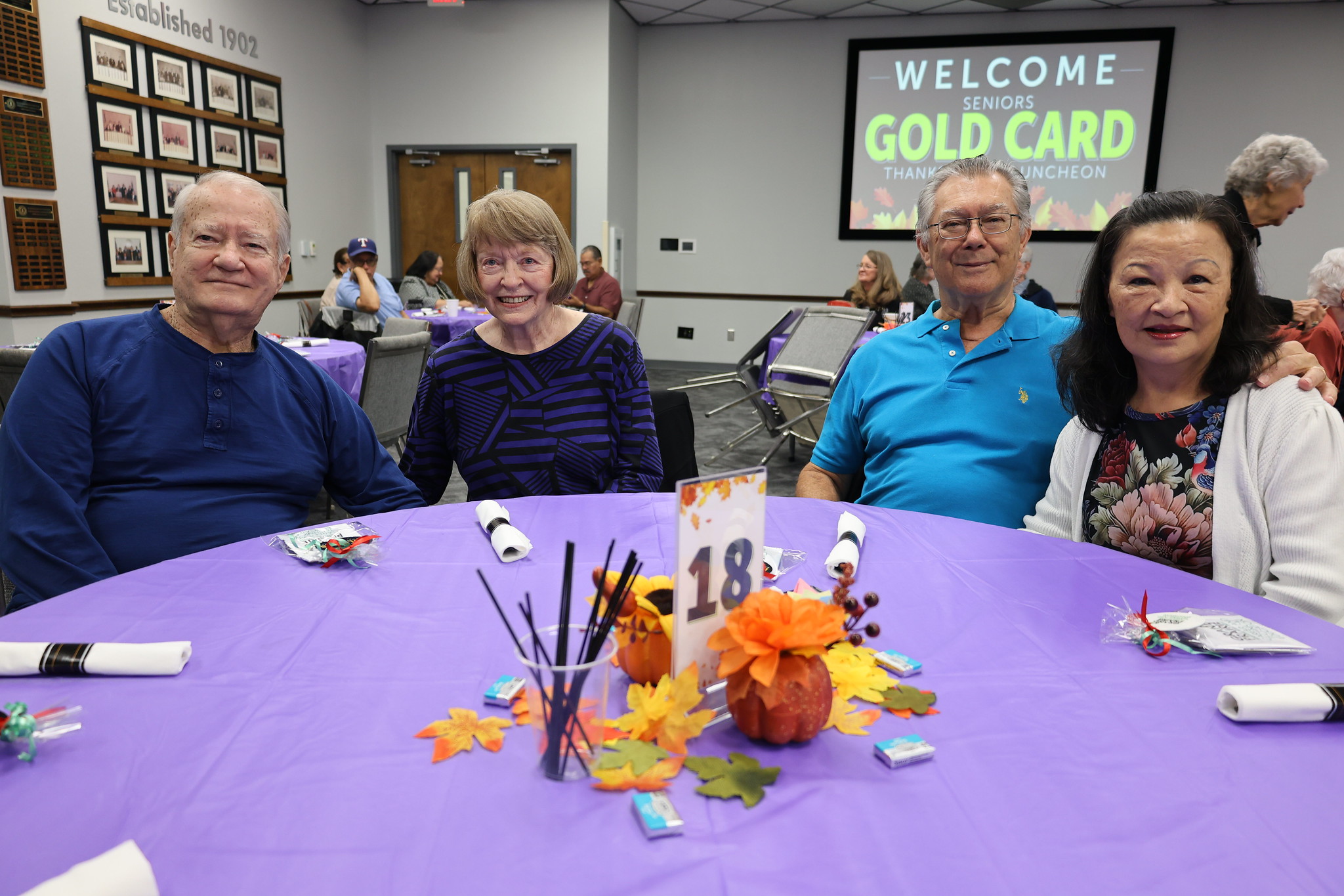 The image shows a group of four older adults sitting around a table, with a colorful autumn-themed centerpiece in the foreground. The background includes a "Welcome Gold Card" sign, indicating this is likely a gathering or event for senior citizens.