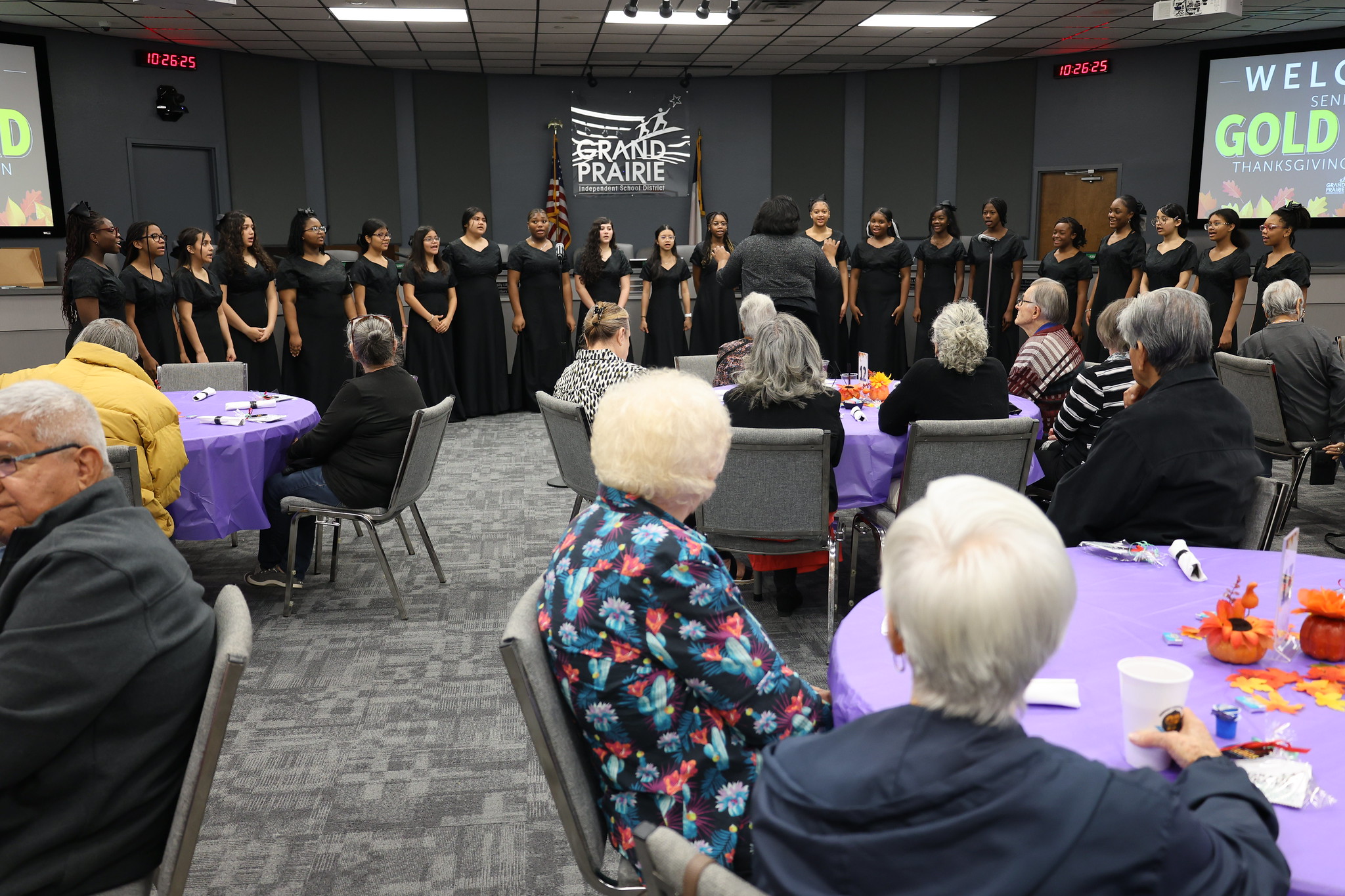 A group of people in black outfits are standing in a room, facing an audience seated at tables in the foreground.