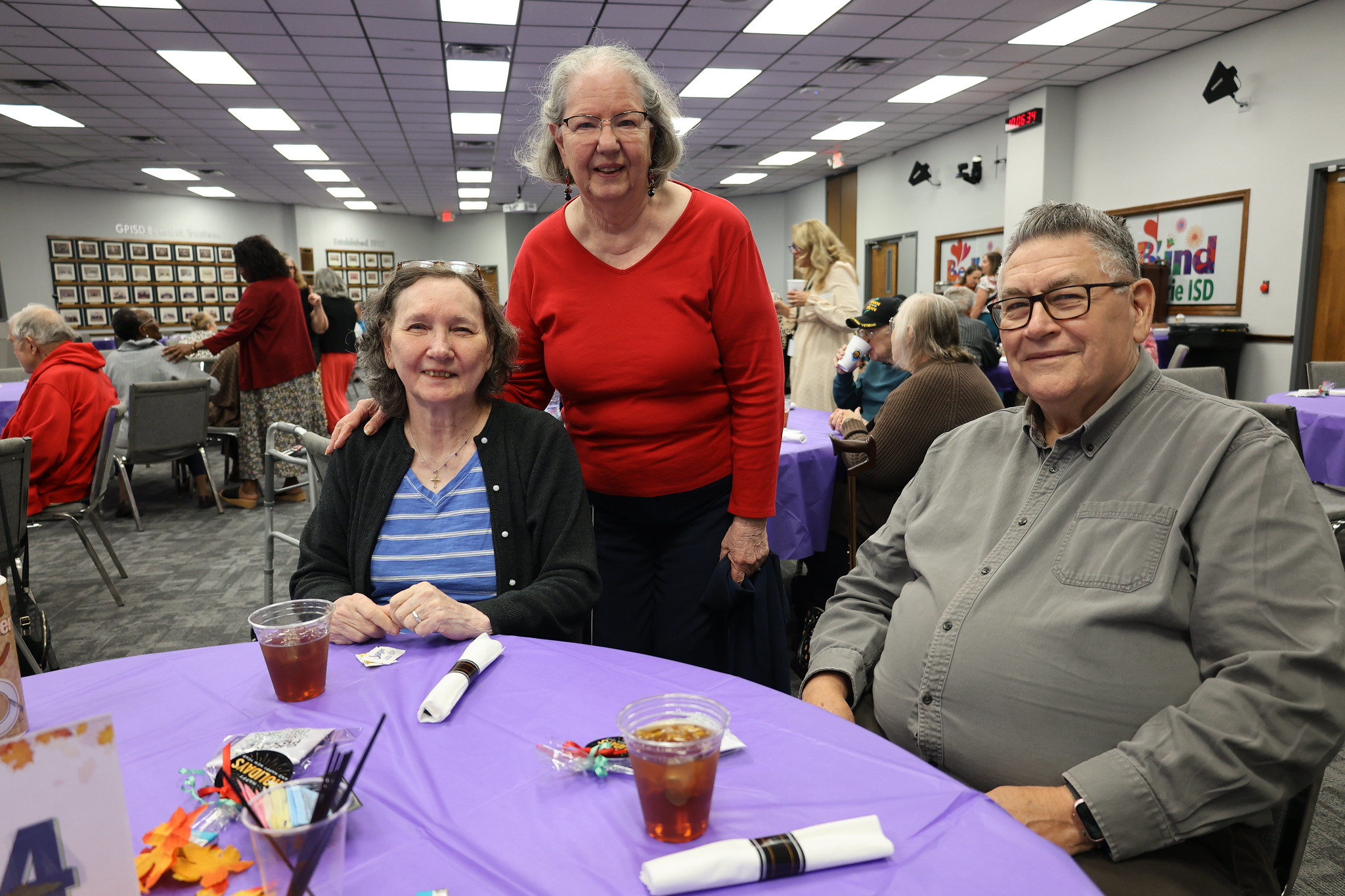 The image shows three older adults, two women and one man, sitting at a table in what appears to be a community center or similar setting, with various decorations and artwork visible in the background.