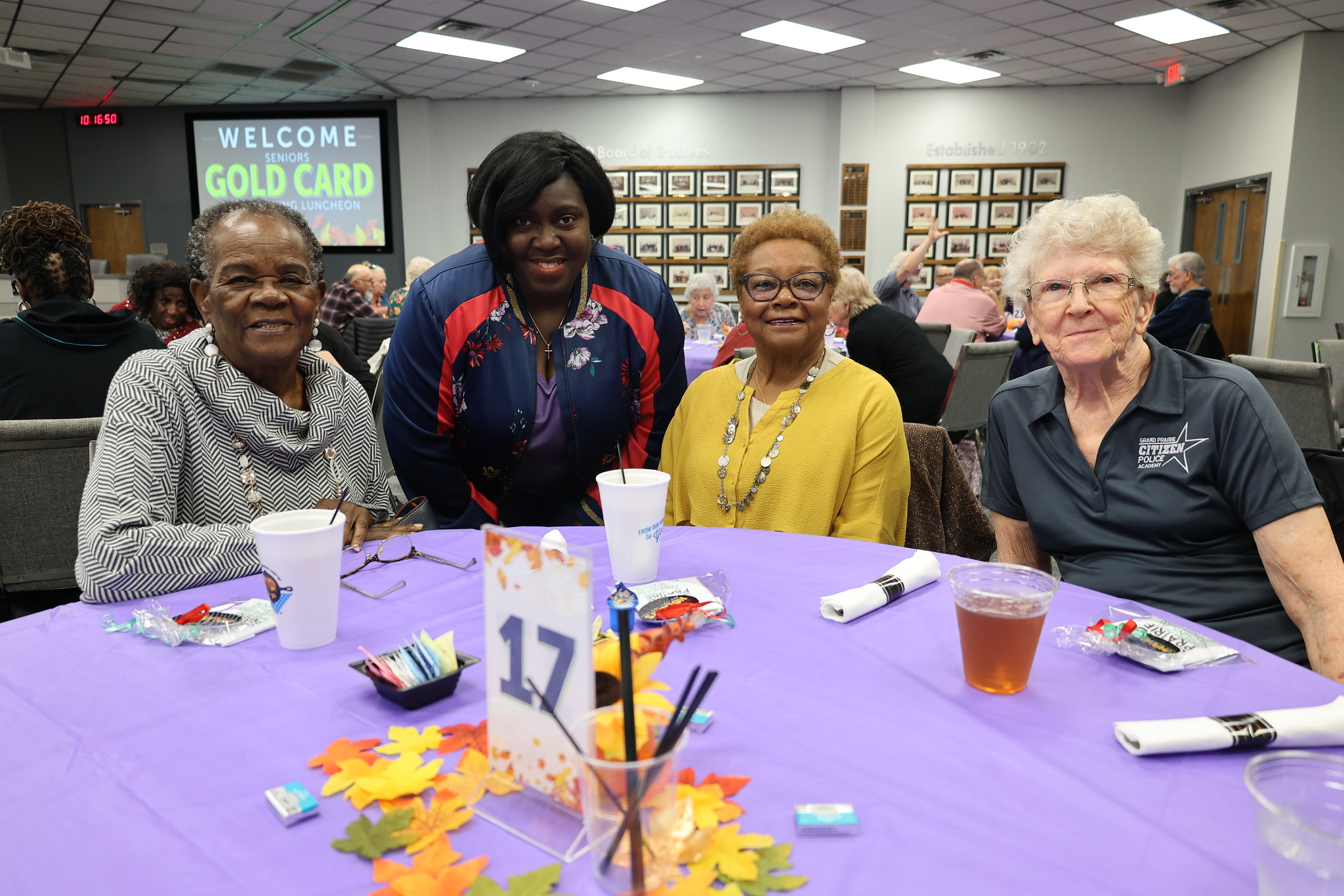 The image shows a group of four older adults sitting at a table, surrounded by decorations and artwork on the walls, indicating a community or social gathering.