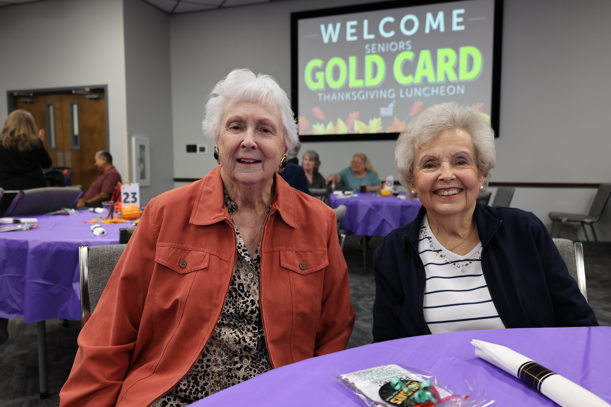 Two elderly women in casual clothing are smiling and standing together in front of a "Welcome Gold Card" sign in what appears to be a community event or gathering space.