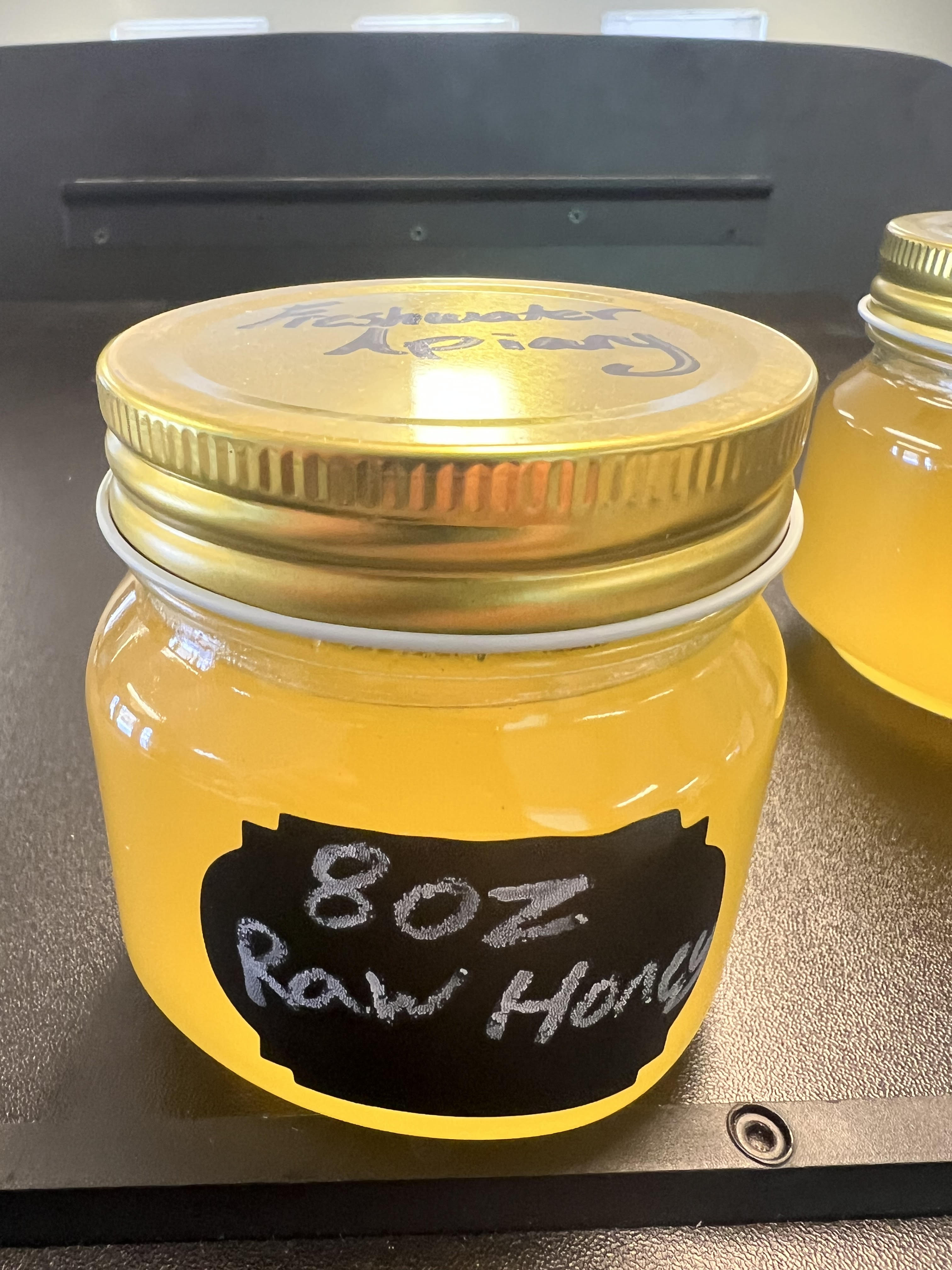 A glass jar filled with a golden-colored liquid, labeled "8oz Raw Honey", sits on a dark surface.