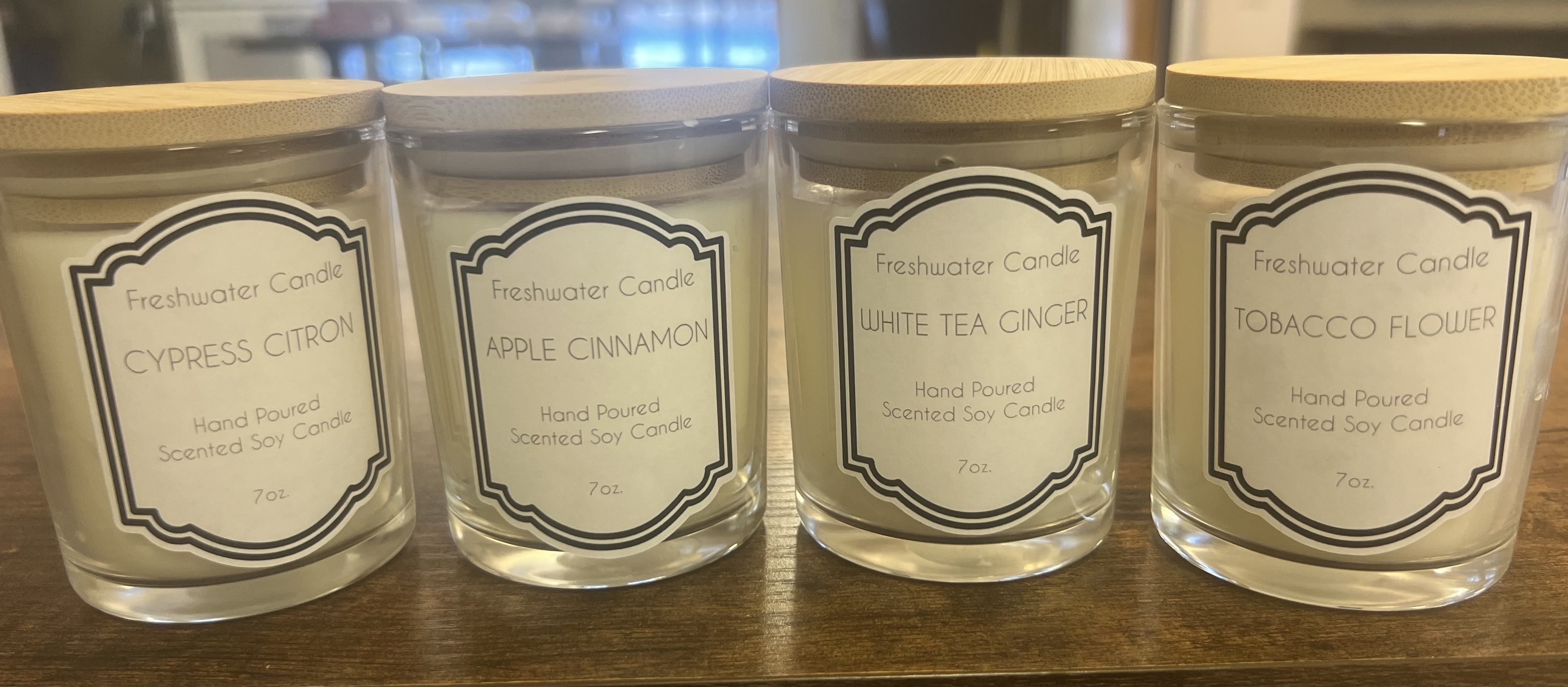 The image shows several glass jars containing various candle scents, such as Cypress Citrus, Apple Cinnamon, White Tea Ginger, and Tobacco Flower, arranged on a wooden surface.