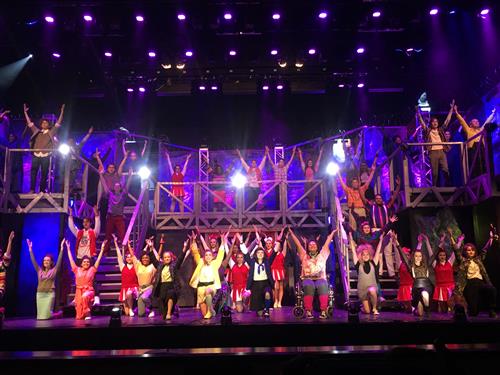 A vibrant and energetic stage performance with a large group of performers in colorful costumes, surrounded by a dynamic lighting setup and a visually striking backdrop.