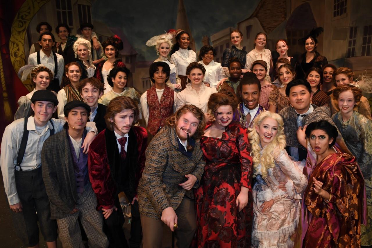 A large group of people, dressed in a variety of colorful and elaborate costumes, posing together in what appears to be a theatrical or performance setting, with ornate backdrops and props visible in the background.