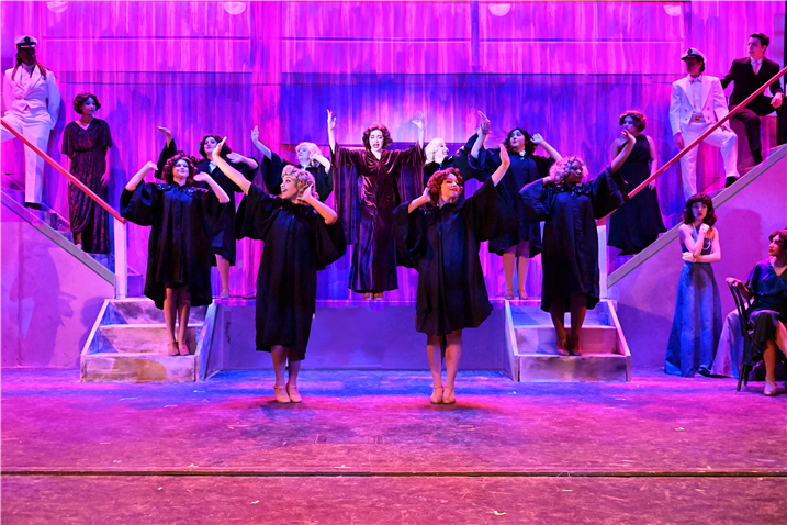 A group of people in black robes stand on a stage with a vibrant purple and pink lighting backdrop, creating a dramatic and theatrical atmosphere.