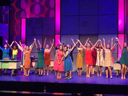 A group of performers in colorful dresses stand on a stage with a vibrant, patterned backdrop and bright, neon-like lighting.