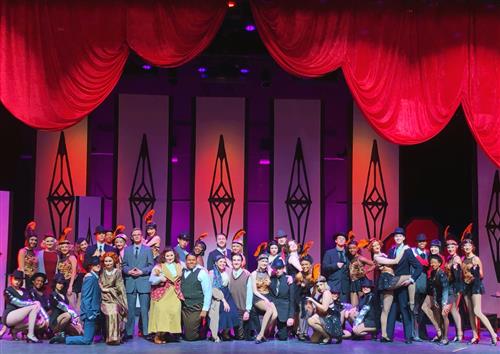 A large group of people, dressed in various formal and theatrical costumes, stand on a stage with a vibrant red curtain and ornate, geometric backdrops.