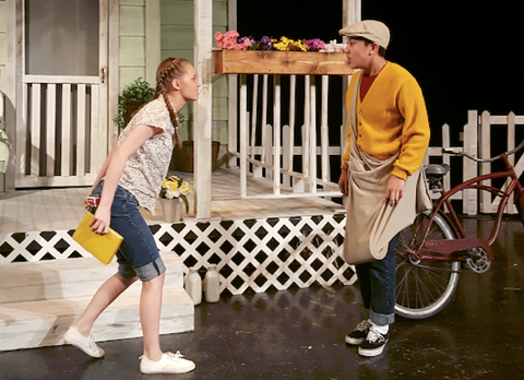 Two people, a man and a woman, are standing on a porch and engaging in conversation. The porch has a lattice railing and a flower box, and the background suggests a residential setting.