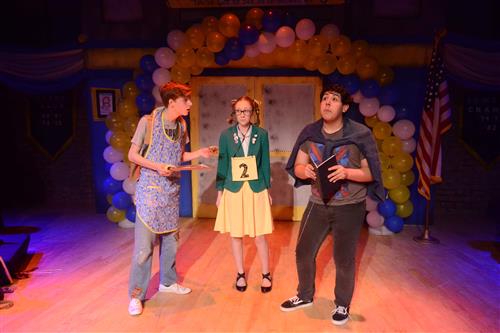 Three individuals, two women and one man, stand on a stage with colorful balloons and decorations in the background, suggesting a performance or event.