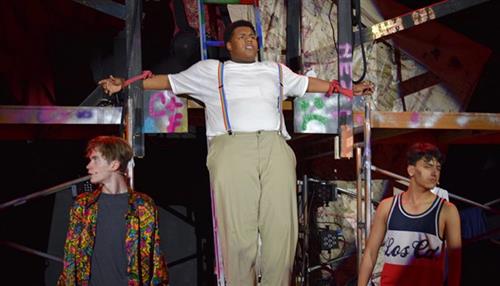 The image shows three individuals on a stage, with one person in the center wearing a white shirt and standing in a confident pose, while the other two individuals are standing on either side of the central figure.