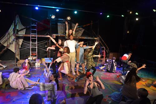 A vibrant and energetic stage performance with dancers in colorful costumes, surrounded by a backdrop of ladders, scaffolding, and lighting fixtures, creating a dynamic and visually striking scene.