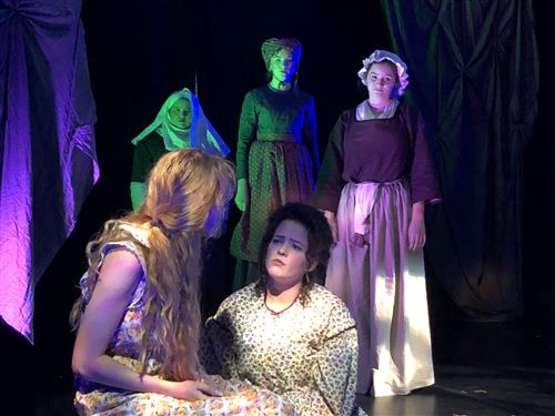 The image depicts a theatrical performance with several costumed actors on stage, including individuals in green and purple costumes, as well as a person in a long dress standing in the background.
