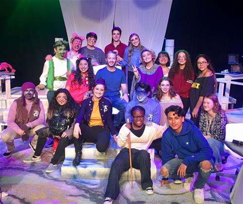 A group of diverse individuals, both seated and standing, posing together on a stage with colorful lighting and backdrops.