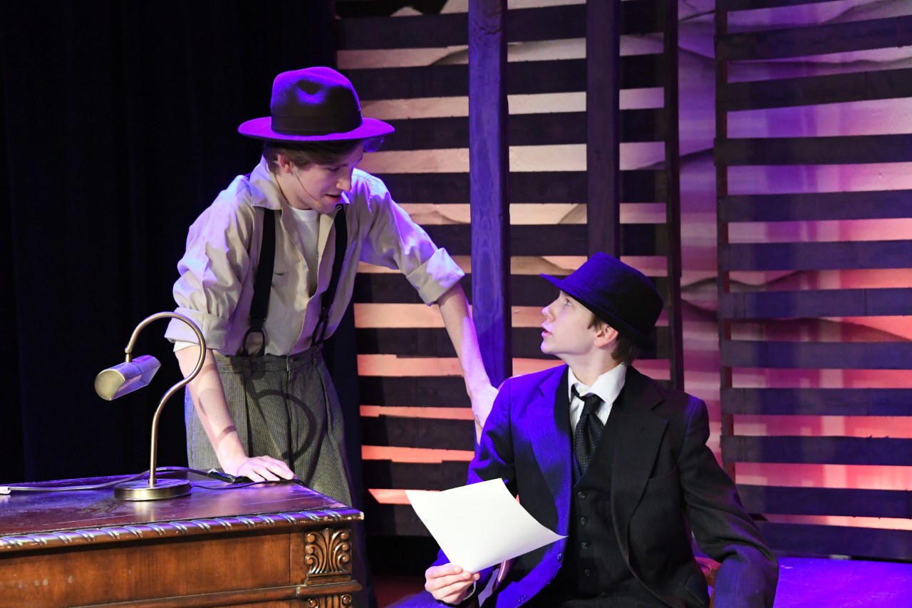 Two individuals, one wearing a colorful hat and the other in a suit, stand on a stage with a wooden piano-like instrument in the foreground, surrounded by a vibrant, patterned backdrop.