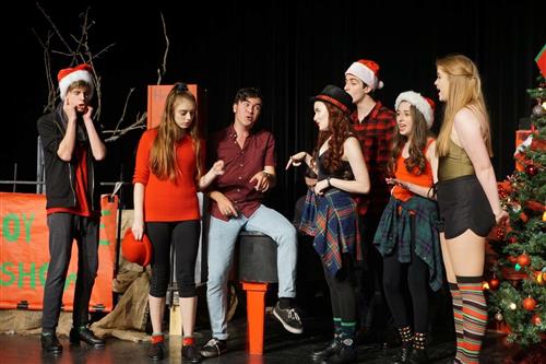 A group of young people dressed in festive attire, including Santa hats and holiday-themed outfits, standing together in front of a Christmas-themed backdrop with decorations and lights.