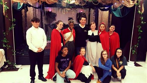 A group of people, some wearing red and others in casual attire, are gathered in front of a decorated backdrop with hanging flowers and greenery.