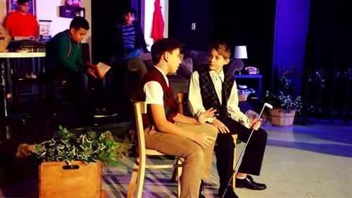 The image shows several individuals seated in a dimly lit room, with various decorative elements and furniture visible in the background.
