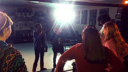 A dimly lit room with several people standing and conversing, with posters and artwork visible on the walls in the background.