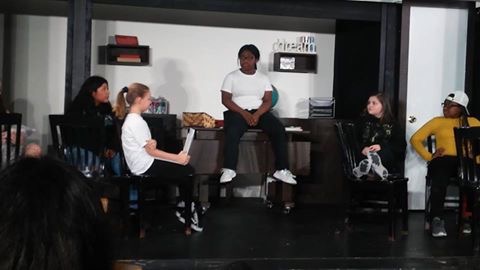 The image shows a group of people gathered in what appears to be a classroom or studio setting, with various shelves and equipment visible in the background.