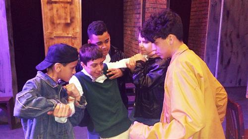 The image shows a group of young men gathered together, some wearing dark-colored clothing and others in lighter shades, standing in what appears to be a dimly lit indoor setting with wooden walls and doors visible in the background.