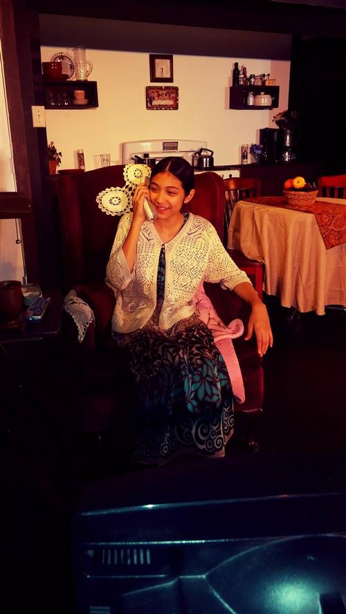 A young woman with a warm smile is seated in a cozy living room setting, surrounded by shelves and decor.