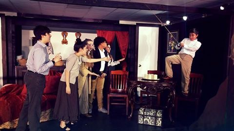 A group of people, dressed in formal attire, are gathered in what appears to be a stage or performance setting, with various props and equipment visible in the background.