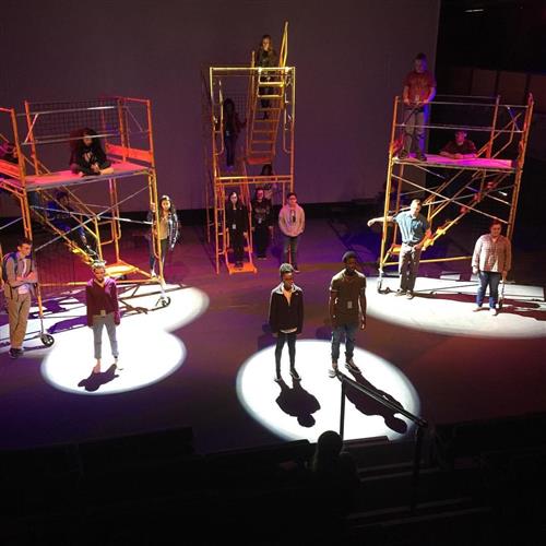 The image depicts a theatrical stage with various props and structures, including scaffolding, ladders, and musical instruments. The stage is illuminated with colorful lighting, creating a dynamic and visually striking scene.
