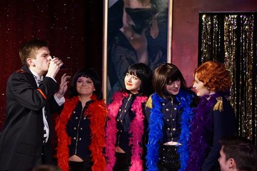 A group of people wearing colorful, feathered costumes stand in front of a dark, curtained backdrop, creating a vibrant and theatrical scene.