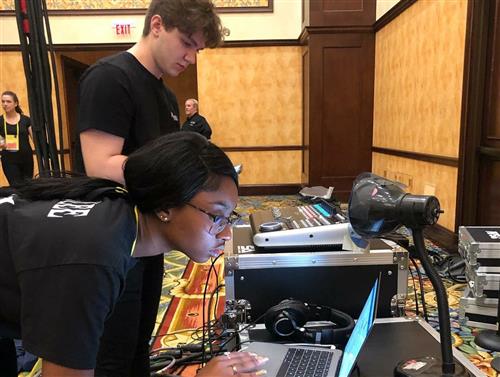 Two individuals, one wearing a black shirt and the other wearing a colorful shirt, are working on a computer setup with various electronic equipment in the background.
