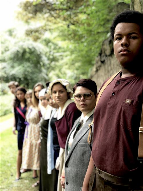 A group of people, including a person in the foreground wearing a burgundy shirt, standing in a wooded area with lush greenery in the background.
