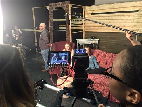 The image shows a film or television production set, with various equipment and crew members visible in the background, while a person is seated in the foreground, likely operating a camera or other filming device.